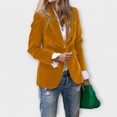 Person wearing a mustard yellow blazer, white shirt, blue jeans, and holding a green handbag on a light gray background